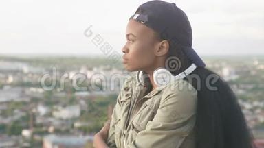 戴着帽子戴着耳机的可爱的非洲裔美国女孩若有所思地看着别处。模糊的城市景观背景